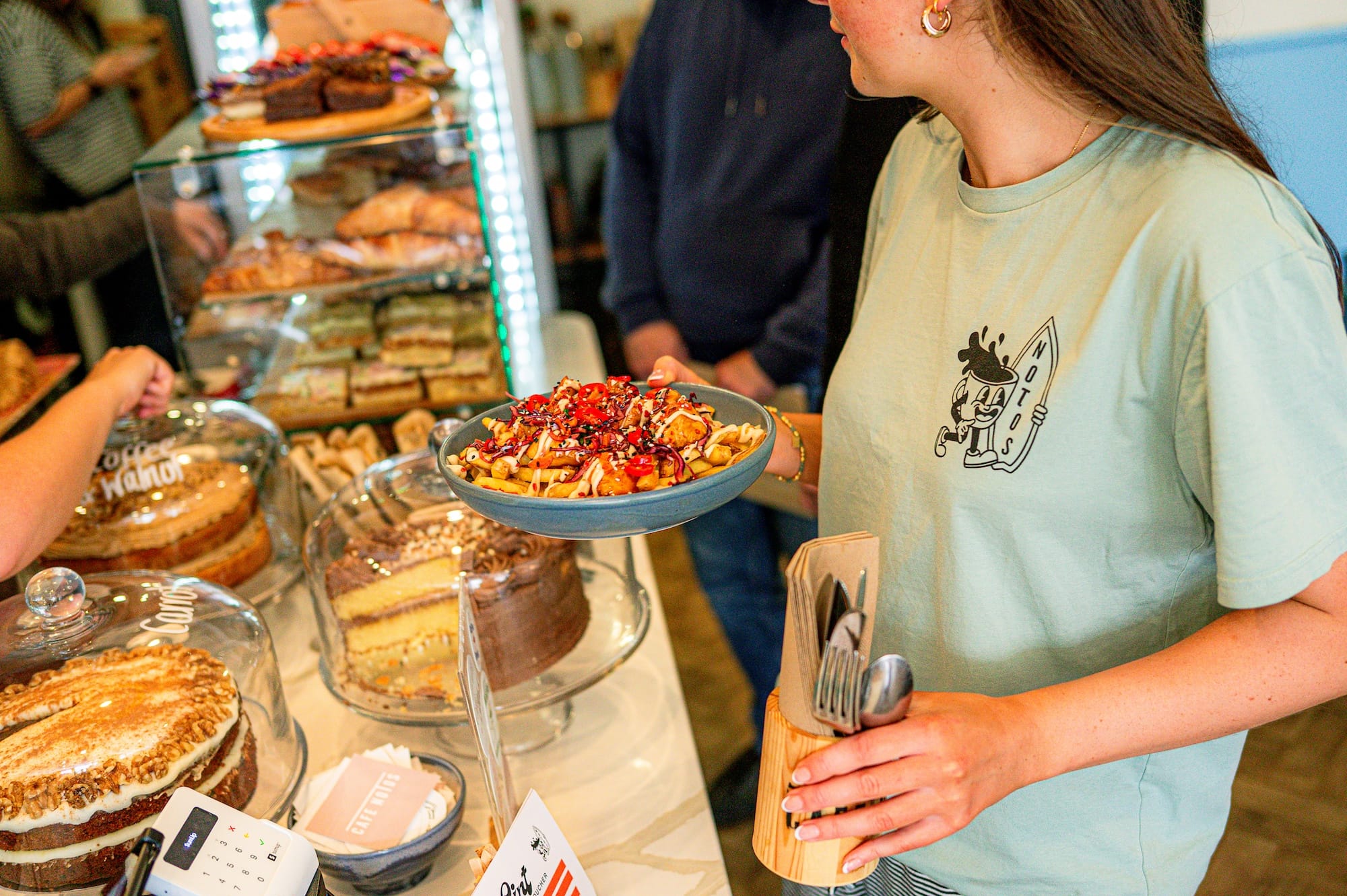A woman holding a plate of loaded fries stands near a display of cakes and pastries in a café.