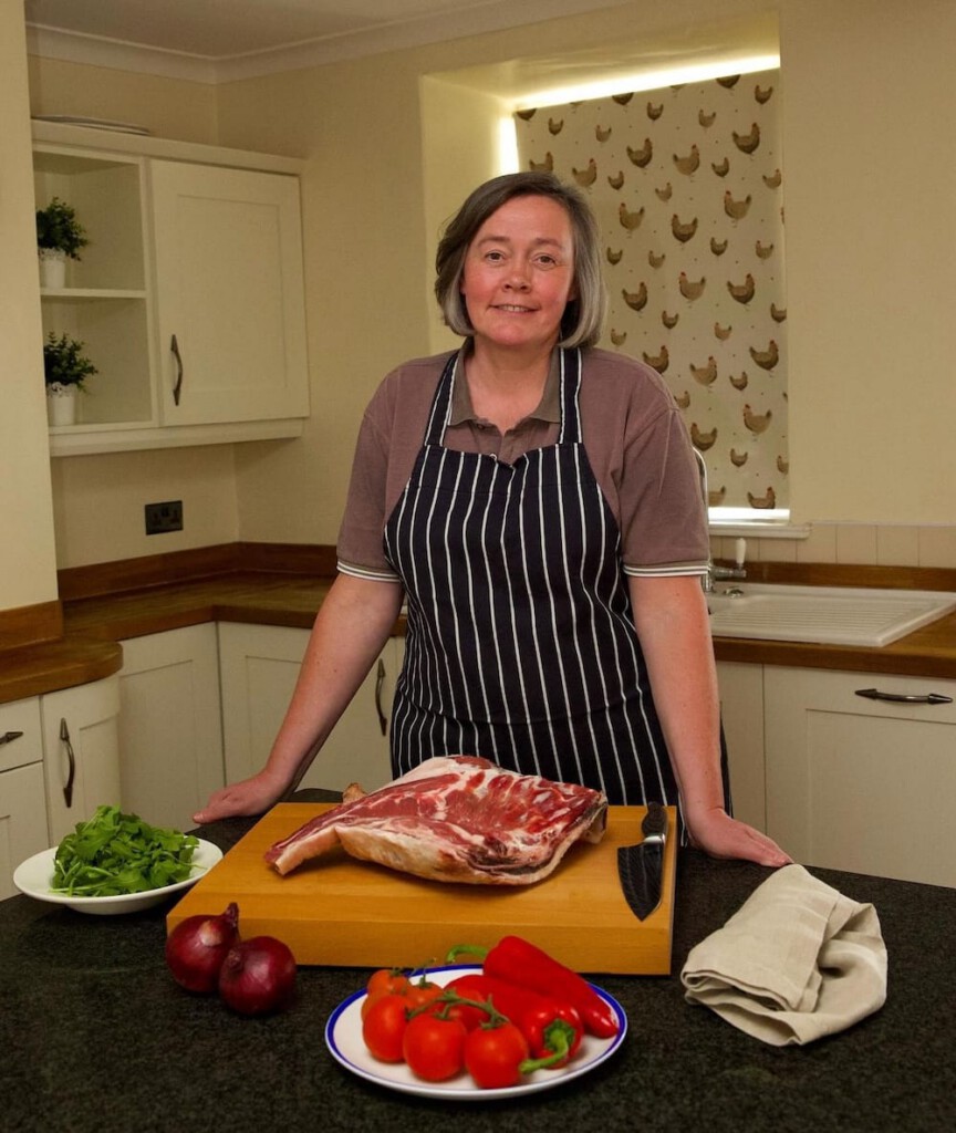 A person in an apron stands in a kitchen behind a counter with a raw rack of lamb, tomatoes, peppers, onions, and a bowl of greens.