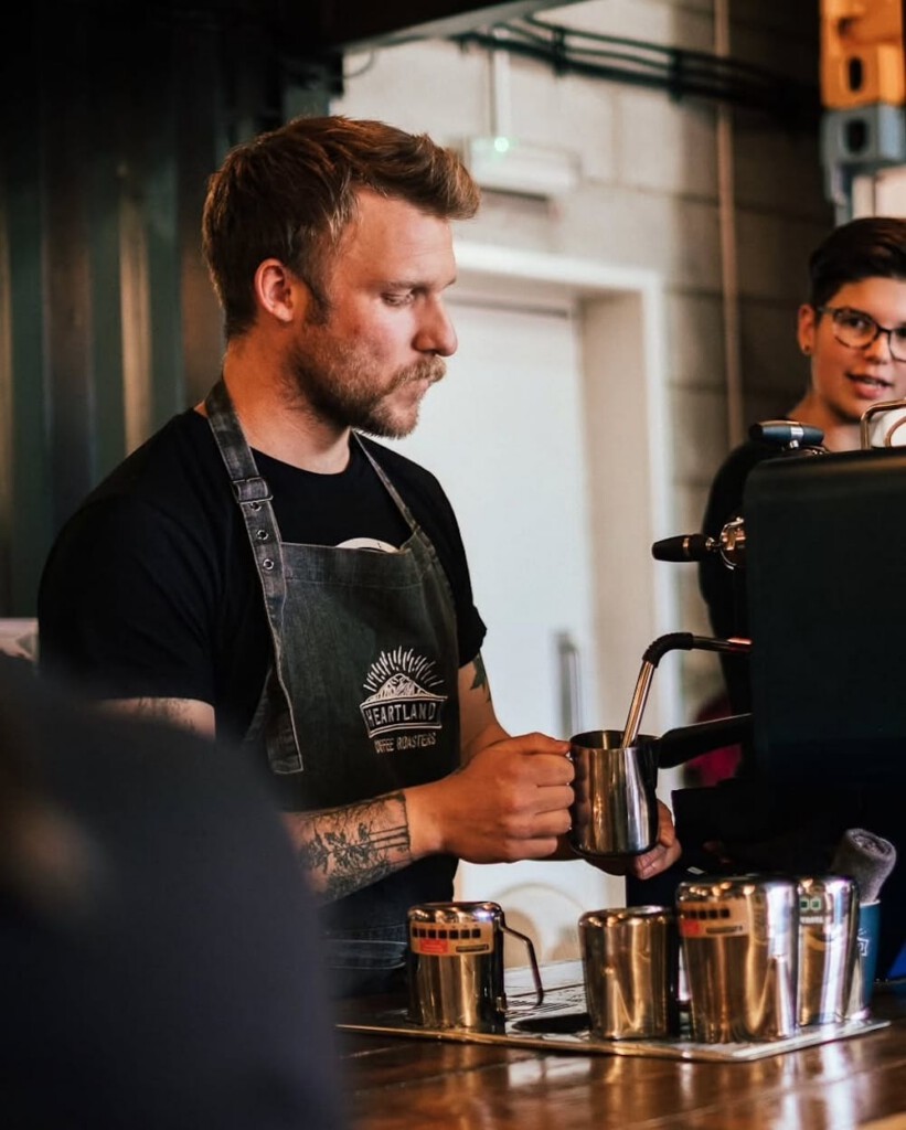 A barista steaming milk with an espresso machine at a coffee shop counter, wearing a Heartland Coffee Roasters apron. Another person stands in the background.