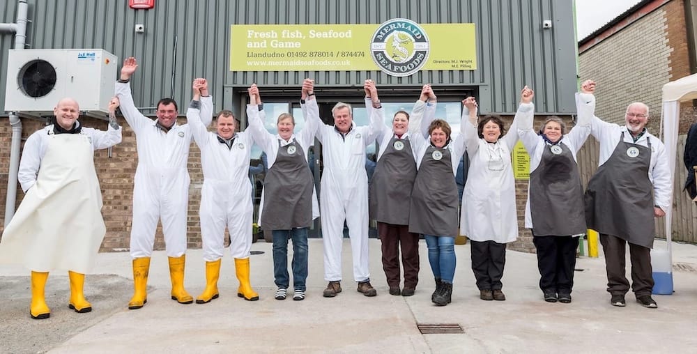 A group of eleven people in uniforms and aprons stand in front of a seafood shop, holding hands and raising their arms in celebration.