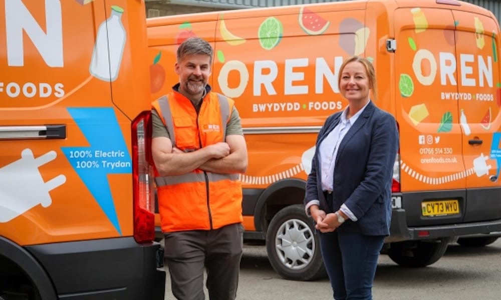 Two people stand smiling in front of two orange Oren Foods delivery vans, one wearing an orange safety vest and the other dressed in business attire.