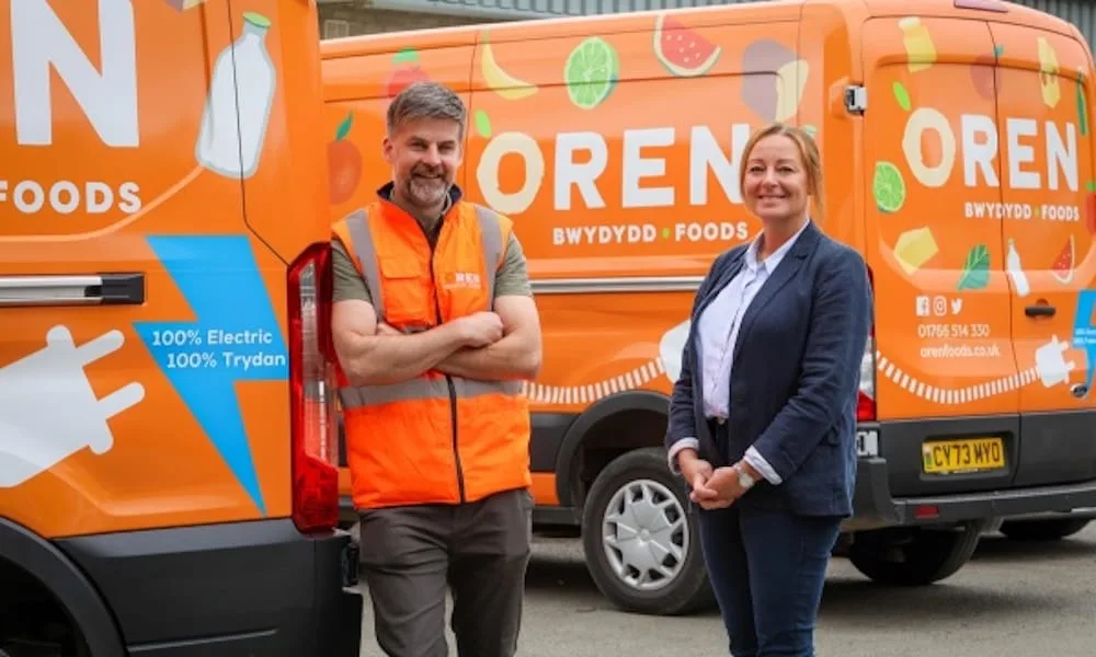 Two people stand smiling in front of two orange Oren Foods delivery vans, one wearing an orange safety vest and the other dressed in business attire.