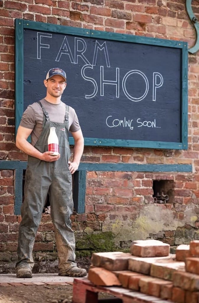 Mae dyn mewn oferôls yn dal potel laeth ac yn sefyll o flaen arwydd bwrdd du ar wal frics sy'n darllen Siop Fferm Yn Dod yn Fuan.