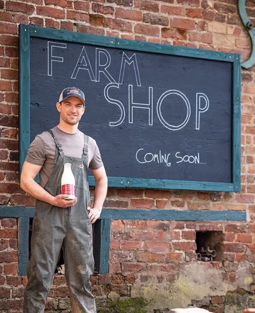 A man in overalls stands in front of a brick wall with a chalkboard sign reading FARM SHOP Coming Soon, holding a bottle of milk.