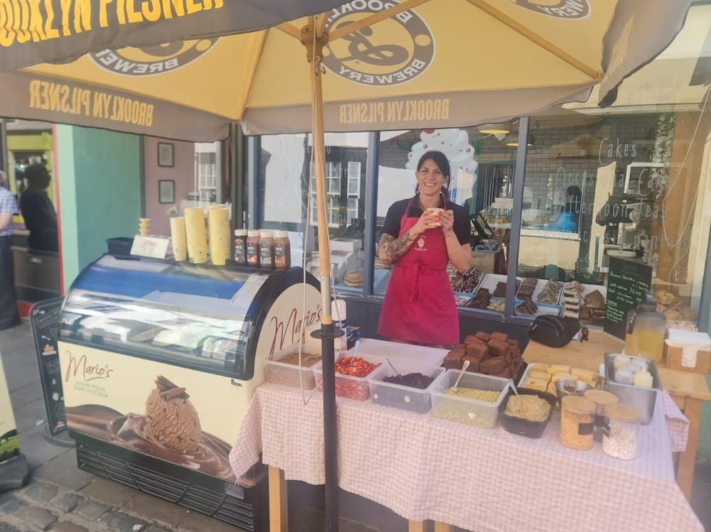 A vendor in a red apron stands behind an outdoor dessert stall with various toppings and baked goods, under a branded umbrella next to an ice cream freezer.
