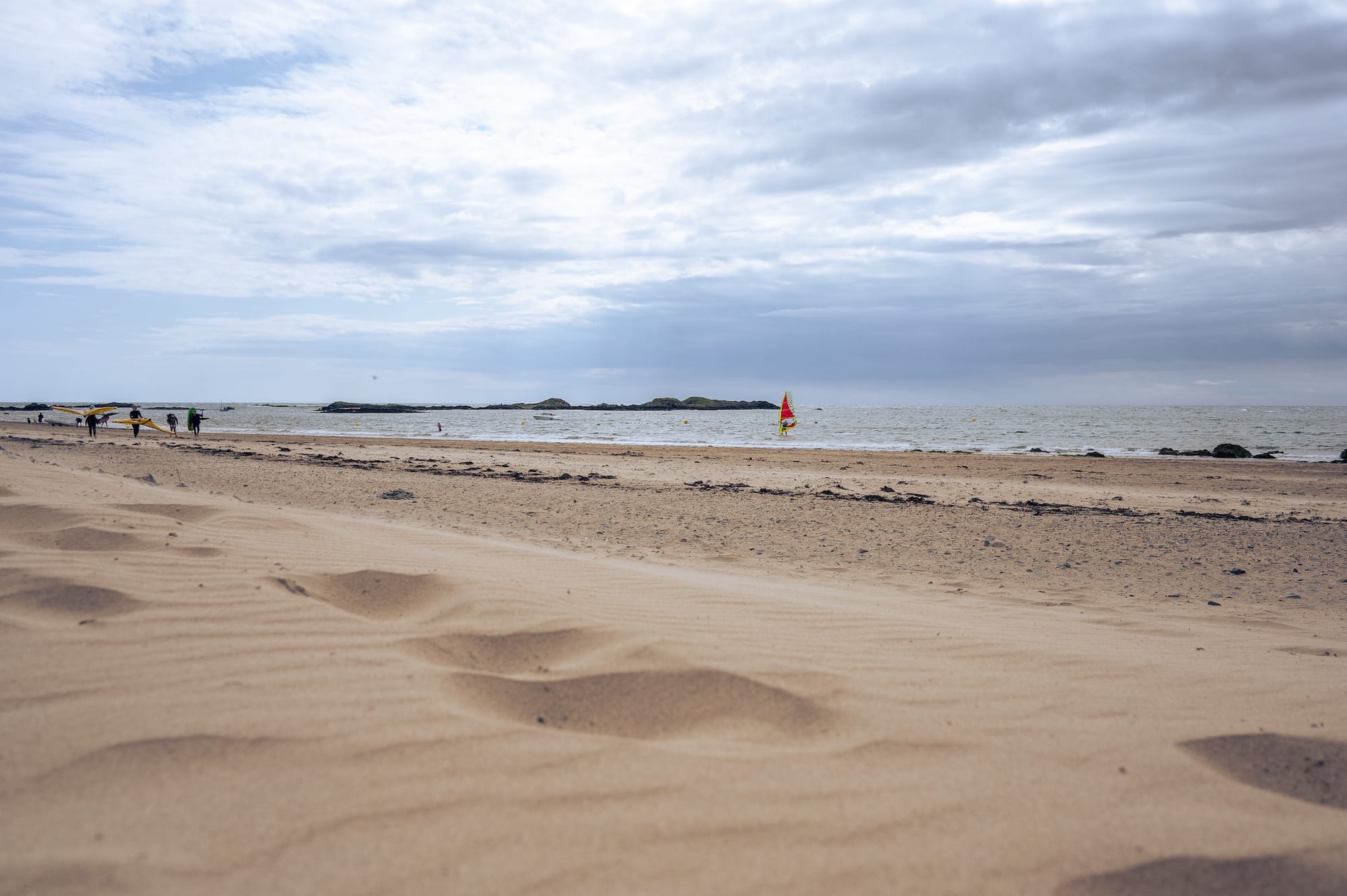 Wide sandy beach with distant footprints in the foreground, a few people near the water, and a windsurfer on the sea under a cloudy sky.