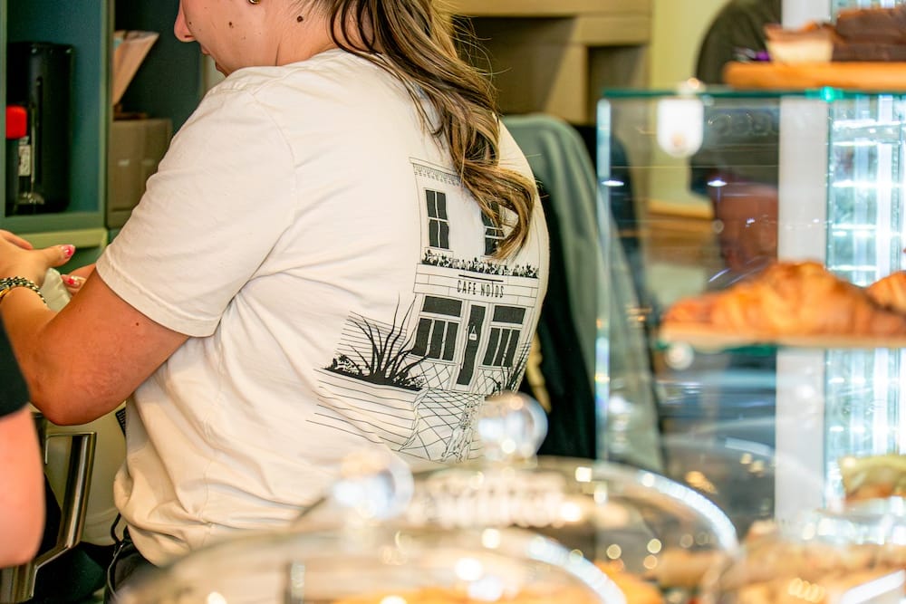 A person wearing a white t-shirt with a building graphic stands at a counter in a bakery or café, with pastries visible in the foreground.