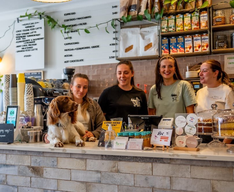 Four women stand behind a cafe counter with a dog, surrounded by coffee machines, menus, and various products on display.