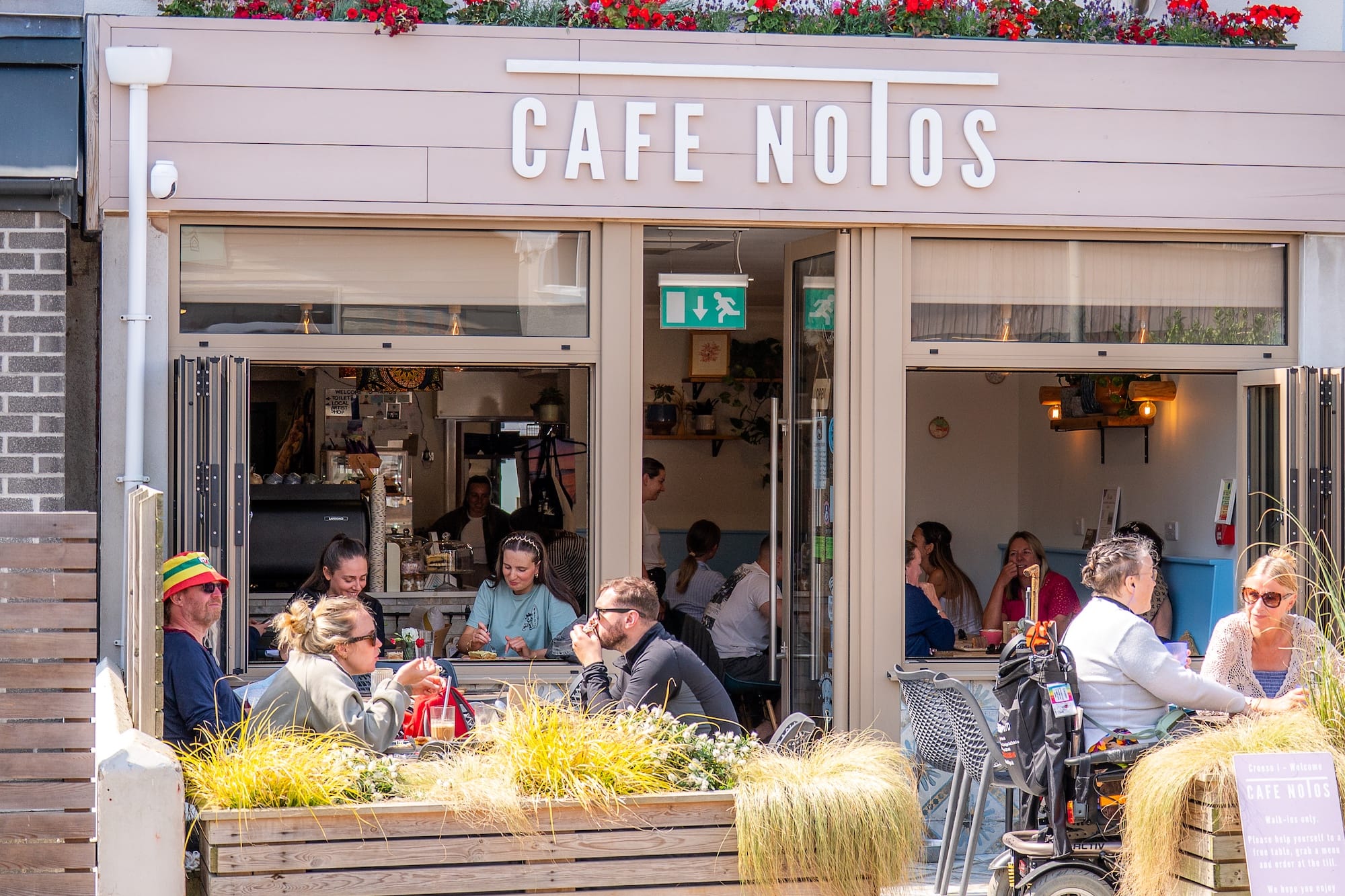 People sit and talk at outdoor and indoor tables at Cafe Notos on a sunny day, with flowers and plants decorating the patio area.