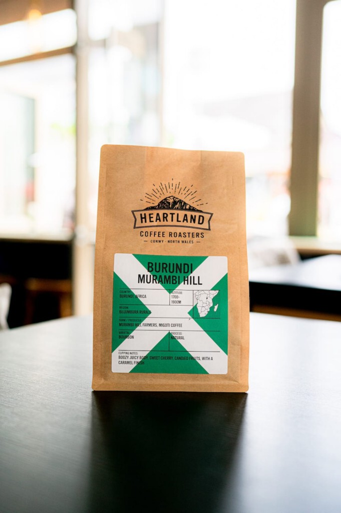 A brown bag of Heartland Coffee Roasters Burundi Murambi Hill coffee sits on a dark table, displaying its green and white label with product information in a well-lit room.