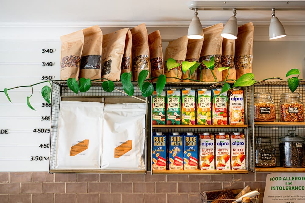 Shelves displaying various plant-based milks, cereals, granola, and paper bags, with a plant draped across the top.