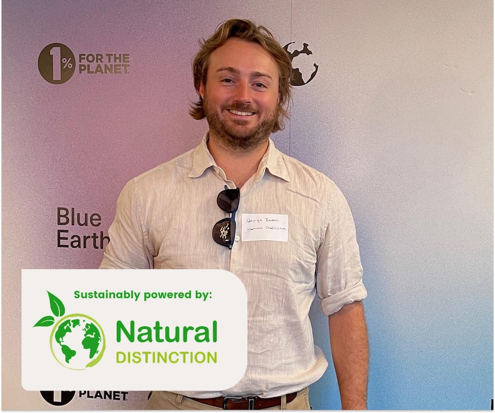 A man with blond hair and a beard stands smiling in front of a wall with environmental logos, wearing a light shirt and a name tag with sunglasses clipped to it. A logo with 'Sustainably Powered by Natural Distinction' is in the bottom left corner.