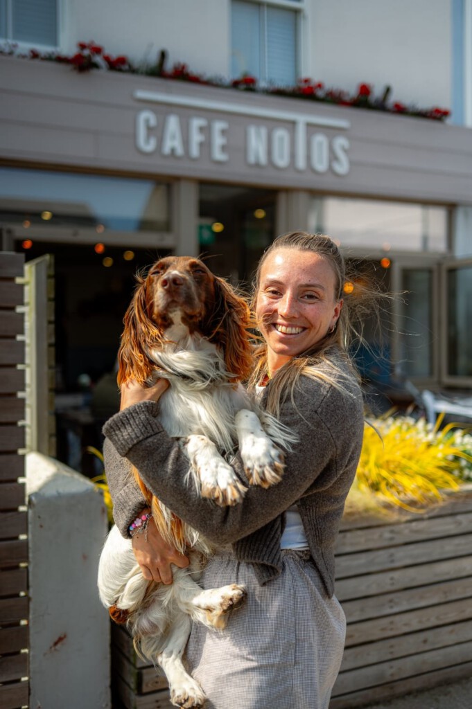 A woman smiling and holding a dog in front of a building with a sign that reads CAFE NOTOS.