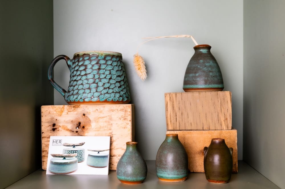 Assorted ceramic vases and a textured mug are arranged on wooden blocks and shelves, with a postcard featuring similar ceramics displayed in the lower left.