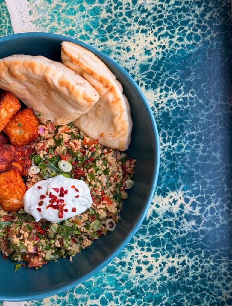 Close up image of a tabbouleh bowl, with halloumi, pitta bread, salad and a white dip