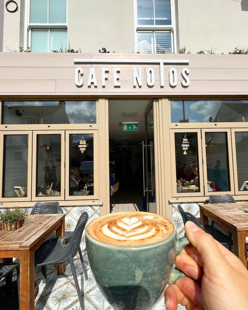 Someone holds a cup of coffee with latte art infront of a cafe shop front in the sun.