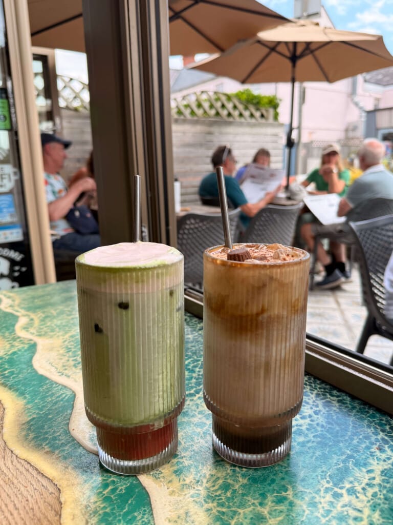 Two cold drinks in a glass, Reese's Iced Mocha and Kiwi & Strawberry Iced Matcha. A busy cafe environment in the background.