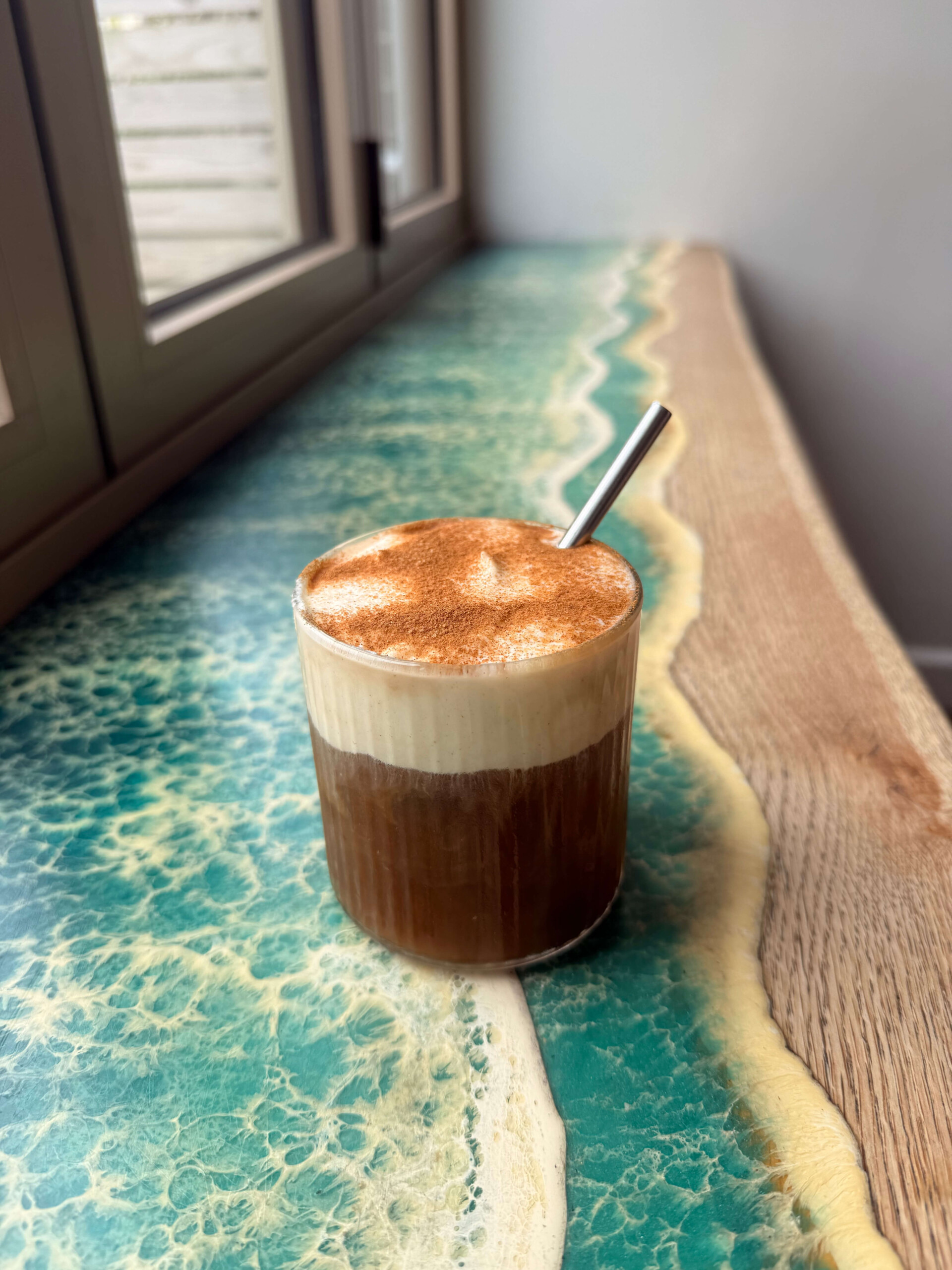 A cold drink glass of Cinnamon Toast Cold Brew on a window shelf.