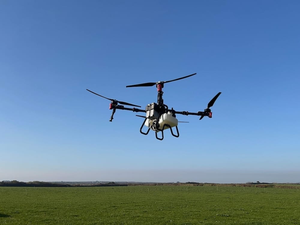 A quadcopter drone with a white body and black arms flies above a grassy field under a clear blue sky.