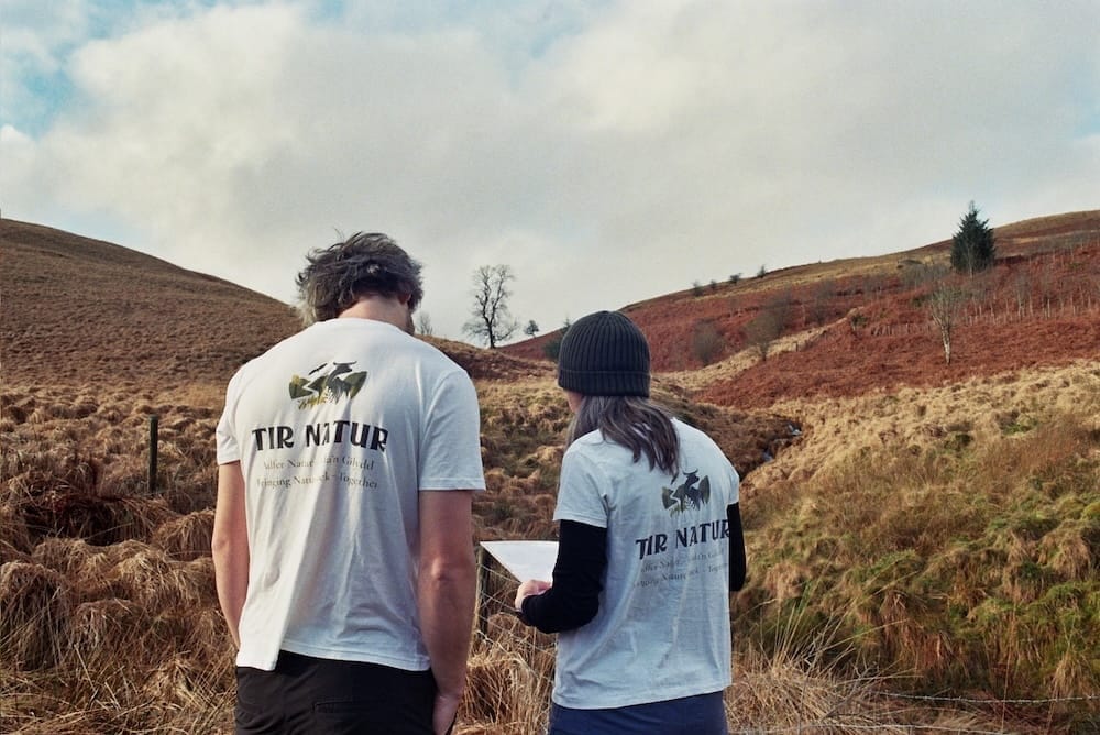 Two people wearing Tir Natur t-shirts stand outdoors in a grassy, hilly landscape, looking at a map under a cloudy sky—an inspiring snapshot of rewilding Wales in action.