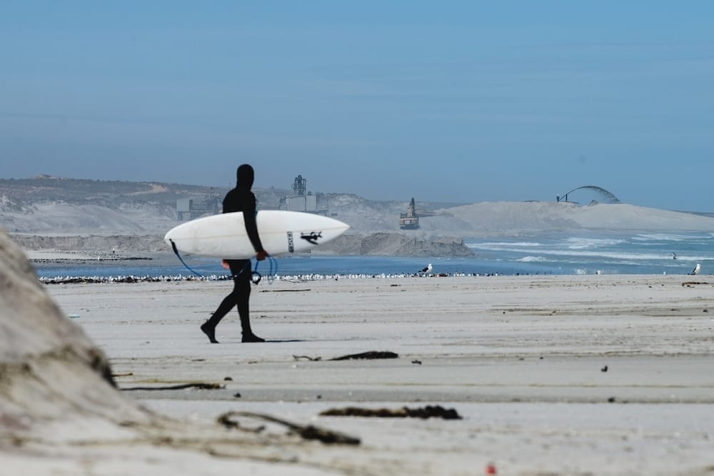 A person in a wetsuit carrying a surfboard walks along a sandy beach with seabirds nearby and industrial structures in the background, reflecting the need to protect the West Coast of South Africa and similar coastal areas like Wales.
