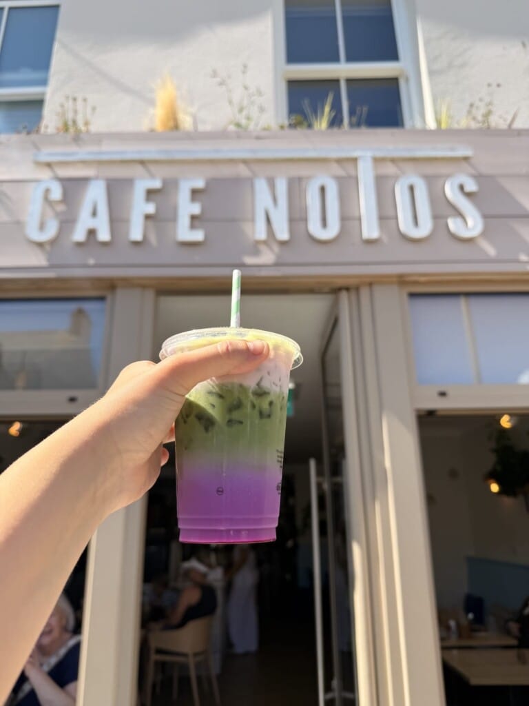 A hand holds a clear cup of iced green and purple layered drink with a straw in front of a cafe entrance with CAFE NOTOS sign.