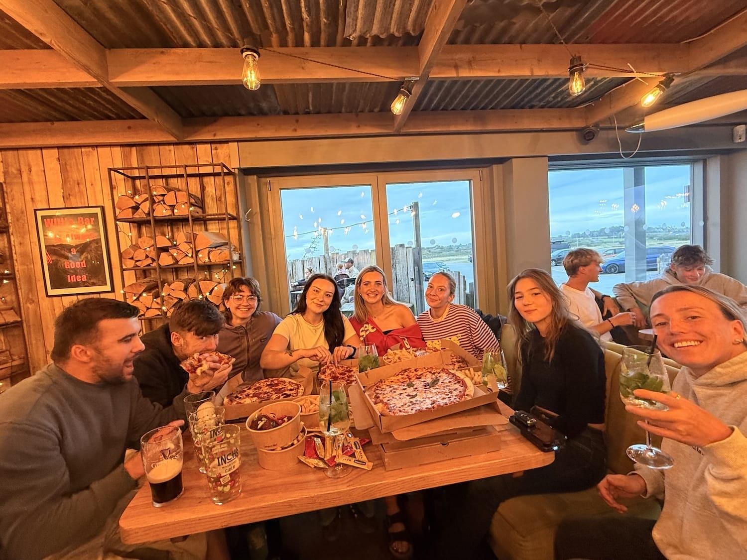 A group of people sit around a table with pizza and drinks inside a wooden restaurant, smiling at the camera.