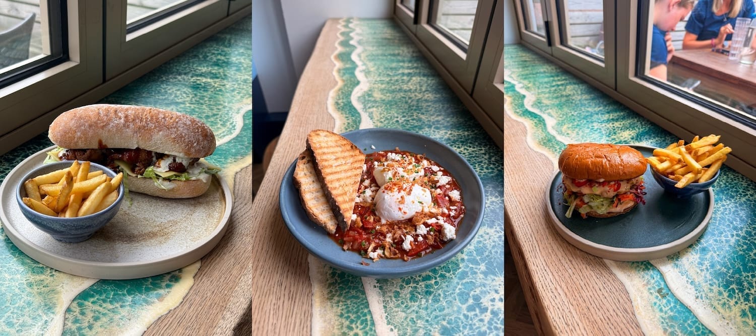 Three dishes on a river-patterned table: a sandwich with fries, poached eggs with toast, and a burger with fries, each near a window.