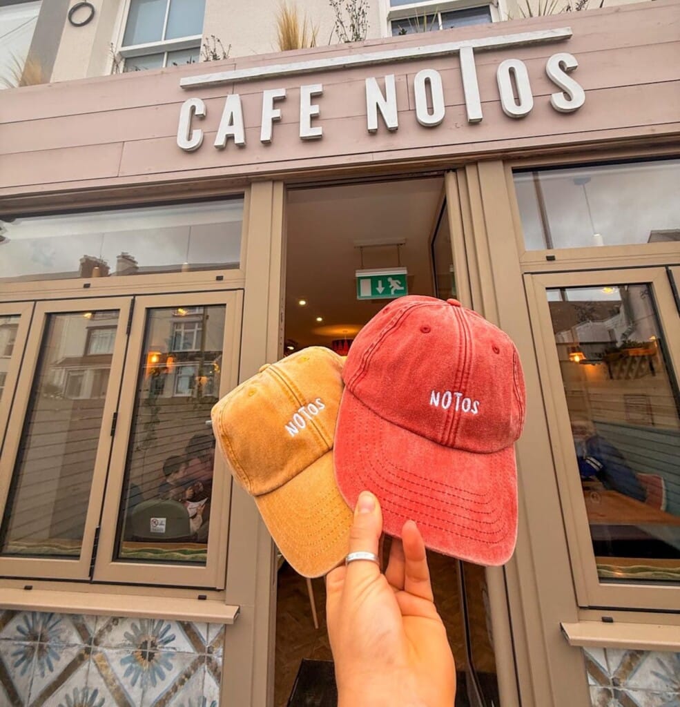 A hand holds two caps, one yellow and one red, both labeled NOTOS, in front of the exterior of a café with a sign reading CAFE NOTOS.